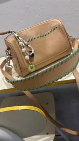 Cartera de cuero color beige con doble correa, una de cuero fina y otra ancha con trenzado en contraste de color verde.