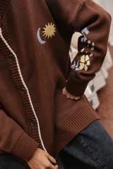 Saco tejido color chocolate con diseño inspirado en la granada, con flores y soles bordados. Presenta cierre frontal con botones, cuello redondo y puños acanalados.