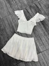 Conjunto blanco de top y short pollera. El top es corto, con escote en V, breteles anchos y volados en los hombros. El short pollera es corto, de corte evasé y cintura alta.