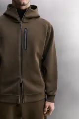 Sudadera con capucha y cierre de cremallera frontal, color negro, confeccionada en tejido técnico compacto de algodón elástico. Presenta cuello subido, bolsillos frontales con cremallera y acabado termofijado, y puños y bajo en rib del mismo tejido.