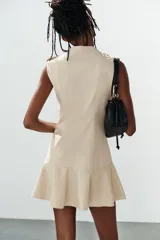 Vestido mini color beige sin mangas, con escote en V, doble botonadura frontal de botones oscuros y bajo con volante amplio. Presenta trabillas en los hombros y falsos bolsillos con solapa.