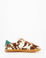 Sneakers de estilo urbano con estampado animal print tipo vaca, confeccionadas en material con textura de pelo. Presentan cordones en tono rosado, detalle de talón en color verde y suela de goma color caramelo.
