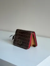 Billetera pequeña de cuero color marrón con textura símil cocodrilo, cierre perimetral en tono rojo y detalles interiores en color tostado. Cuenta con un aplique metálico circular en la parte frontal.