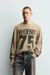 Sudadera de corte relajado confeccionada en tejido con textura waffle, color beige con efecto lavado. Presenta cuello redondo, manga larga con franjas laterales oscuras y estampado frontal con texto y número en contraste.