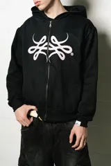 Hoodie con cierre frontal, color negro, de corte regular fit. Presenta un estampado frontal grande en blanco con un diseño abstracto y estilizado, similar a un monograma o símbolo.