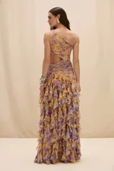 Vestido largo de un solo hombro con estampado floral en tonos amarillos y violetas. Presenta un diseño con aberturas laterales en la cintura, cuerpo drapeado y falda con volados en cascada y tajo lateral profundo.