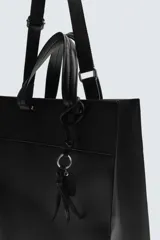 Bolso de hombro liso color negro, con cierre de imán en el compartimento principal y bolsillo interior con cremallera. Incluye un charm decorativo, dos asas de mano y un asa de hombro ajustable y extraíble.