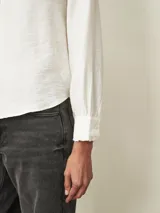 Camisa de manga larga en color blanco, con cuello mao y detalle de volados sutiles en el borde. Presenta cierre frontal oculto con botones y puños con volados.