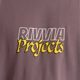 Remera violeta de manga corta con cuello redondo y estampado frontal con el logo de la marca Rivvia en amarillo.