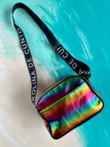 Bandolera Kaira de la línea Ottenta de Carolina De Cunto, confeccionada en material metalizado con acabado multicolor y efecto tornasolado. Cuenta con un bolsillo frontal con broche imantado, cierre superior y una correa larga y ajustable de tela negra con el nombre de la marca en letras blancas.