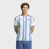 Camiseta de fútbol Adidas de la selección argentina, modelo Primera Equipación 26. Diseño a rayas verticales blancas y celestes con un efecto degradado en las franjas celestes. Presenta cuello redondo con ribete azul oscuro y tres franjas de Adidas en los hombros en azul oscuro. El escudo de la AFA y el logo de Adidas son de color dorado.