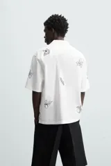 Camisa de manga corta confeccionada en mezcla de algodón y lino, color blanco. Presenta un cuello tipo bowling, calce regular y cierre frontal con botones. Destaca por sus bordados lineales en contraste de color negro con motivos de frutas y hojas distribuidos por toda la prenda.