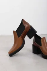 Botín asimétrico de estilo Chelsea color bordó, confeccionado en cuero vacuno, con paneles elásticos laterales negros, puntera cuadrada, suela de plataforma y tacón de bloque cuadrado.