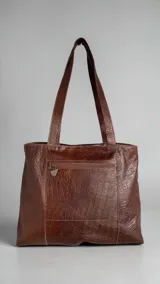 Cartera de cuero natural en color marrón jaspeado, tipo tote bag, con diseño amplio y asas dobles para el hombro. Presenta intervención artística pintada a mano en el frente con detalles metalizados y geométricos, y dije metálico colgante.