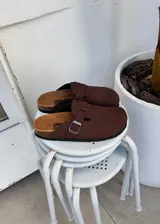 Zuecos de gamuza color marrón chocolate, con diseño cerrado en la parte delantera y tira ajustable con hebilla metálica en el empeine. Presentan plantilla anatómica de corcho y suela de goma.