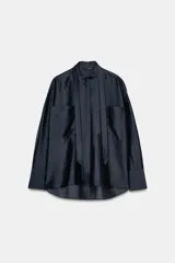 Camisa de corte holgado confeccionada en mezcla de lyocell con acabado satinado. Presenta cuello redondo con lazadas integradas, cierre frontal con botones ocultos bajo solapa, bolsillos de plastrón en el frente y mangas largas con puños anchos. El bajo es asimétrico y redondeado.