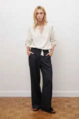 Camisa de color crudo con diseño romántico, cuello redondo con cordón ajustable terminado en borlas, y mangas tres cuartos abullonadas. Presenta detalles de bordados calados verticales en el frente y horizontales en las mangas.