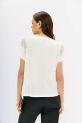 Remera blanca de lino de textura suave y liviana, con calce al cuerpo y mangas cortas. Presenta cadenas de strass decorativas en los hombros.