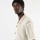Camisa de manga corta confeccionada en tejido de punto fantasía color beige, con cuello camisero, cierre frontal de botones y detalles de líneas oscuras en zigzag.