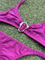 Bikini de tela metalizada color magenta, compuesto por un top con breteles finos y detalle de hebilla metálica en el centro, y una bombacha tipo colaless.