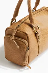 Cartera bowling chica color marrón con textura símil cuero, cierre superior y doble asa corta con argollas metálicas doradas. Cuenta con dos compartimentos laterales con cierre y forro de tafeta.