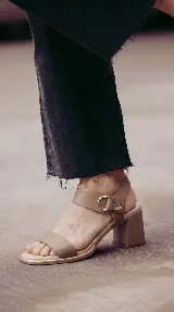 Sandalia de cuero color taupe con taco cuadrado de 6 cm, tira frontal ancha y tira en el tobillo con detalle metálico dorado y hebilla circular.