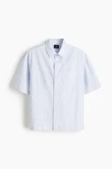Camisa de manga corta en popelina de algodón a rayas verticales celestes y blancas. Modelo boxy con cuello inglés, tapeta clásica, bolsillo superior abierto y canesú en la espalda. Bajo recto con aberturas laterales.