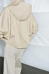 Trench corto color verde militar, con cuello solapa y capucha. Tiene manga larga con trabillas y botón en los puños, bolsillos delanteros y bajo asimétrico. Cierre frontal con botones. El tejido es repelente al agua.