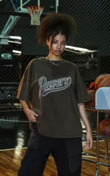 Remera de manga corta y cuello redondo, calce oversize, color marrón oscuro con efecto batik artesanal (lavado ácido). Estampa frontal con la palabra "Ravers" en estilo cursiva con contorno blanco.