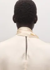 Blusa de seda color blanco marfil, con cuello alto drapeado y detalle de bufanda integrada al cuello. De manga larga con puños ajustados mediante tiras.