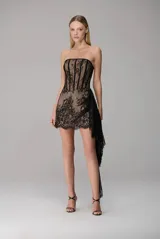 Vestido corto strapless confeccionado en encaje negro sobre forro nude, con diseño estructurado tipo corset en el torso y falda ajustada con detalle de sobrefalda lateral asimétrica y caída larga de encaje.