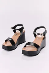 Sandalias de cuero negras con plataforma y tacón de 10 cm. Tienen una tira ancha en la parte delantera y una tira ajustable con hebilla alrededor del tobillo, ambas con detalles de tachas plateadas.
