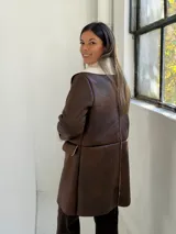 Abrigo de cuero sintético color marrón con interior y solapas de corderito sintético color beige. Presenta cierre frontal con botones, mangas largas con puños vueltos y bolsillos laterales.