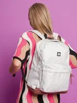 Mochila color gris claro con base marrón, con dos bolsillos frontales y logo cuadrado en la parte superior. Tiene correas negras ajustables y compartimento interno para notebook.