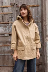 Parka liviana color beige con capucha, cierre frontal con botones, bolsillos delanteros con tapa y ajuste en la cintura. Presenta mangas con puños vueltos y detalle de botones.