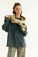 Buzo oversize unisex de color gris verdoso con efecto acid wash, mangas largas y cuello redondo. Presenta costuras frontales decorativas y un pequeño logo bordado en el centro del pecho.