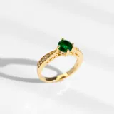 Anillo dorado con cristal verde esmeralda en el centro y pequeños cristales claros alrededor.
