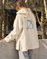 Canguro de felpa color crudo, con capucha, corte amplio, bolsillos delanteros y aberturas laterales en el dobladillo. Presenta un estampado en la espalda con diseño de arco y flecha rodeado de estrellas, y detalles gráficos en las mangas.