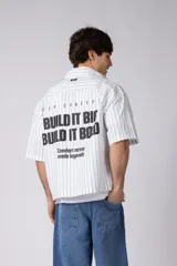 Camisa de manga corta a rayas beige y blancas, de corte boxy y liviana, con cuello camisero, cierre frontal de botones y pequeño texto bordado "BUILD IT BOLD" en el pecho.