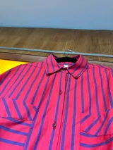 Camisa de corte oversize con diseño de rayas verticales en color rojo y azul. Presenta cuello clásico, cierre frontal con botones, mangas largas con puños abotonados y dos bolsillos frontales con solapa. El diseño tiene un largo asimétrico, siendo más corto en la parte delantera y más largo en la parte trasera.