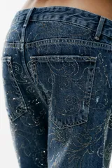 Jeans baggy de tiro medio con cinco bolsillos, trabillas para cinto y cierre frontal con cremallera y botón metálico. Cuenta con detalle de aplicación de brillos en toda la prenda.