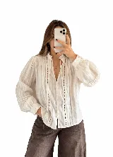 Blusa blanca de estilo romántico, confeccionada en tela liviana con detalles de alforzas y encaje insertion en el frente. Cuenta con cuello alto fruncido con lazo de ajuste, mangas largas abullonadas con puños elásticos y cierre frontal con botones.