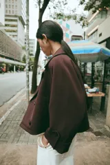 Trench corto de color marrón oscuro, con cuello de solapa, cierre frontal cruzado con botones y mangas tres cuartos con detalle de vuelta, trabilla y botón. Cuenta con bolsillos delanteros de vivo.