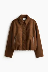 Campera de gamuza color marrón con acabado cepillado suave, cuello clásico, hombros caídos y mangas largas. Cuenta con cierre frontal oculto bajo solapa con botones a presión, bolsillos laterales con cierre y diseño abullonado con elástico interior en el bajo.