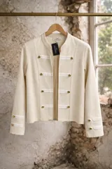 Chaqueta de estilo militar en color beige, confeccionada en tejido de algodón. Presenta cuello mao, cierre frontal con botones dorados y detalles de tiras horizontales en contraste. Cuenta con mangas largas con detalles de botones en los puños.