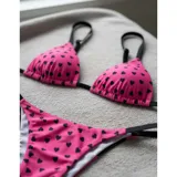 Bikini de dos piezas con estampado de corazones negros sobre fondo fucsia. El top es de estilo triángulo con tiras negras, y la parte inferior es una tanga con tiras laterales negras.