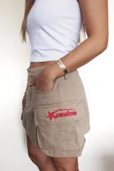 Falda corta estilo cargo color beige, con bolsillos laterales y un parche bordado rojo con una estrella y texto en el bolsillo derecho.