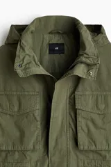 Campera estilo cargo de tejido ripstop, color verde militar, con cuello elevado que incluye capucha oculta. Presenta cierre frontal con solapa rompevientos, cuatro bolsillos delanteros con solapa y botón a presión, puños ajustables y cordón interno en la cintura.