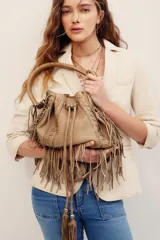 Cartera estilo saco de cuero vacuno marrón, con flecos colgantes, asa tubular rígida y detalles de costuras decorativas. Cuenta con cordón ajustable en la parte superior adornado con borlas y herrajes metálicos.