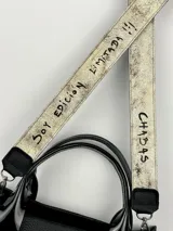 Bolso negro de cuero con detalles charolados, doble asa corta y correa larga de tela con el nombre de la marca.